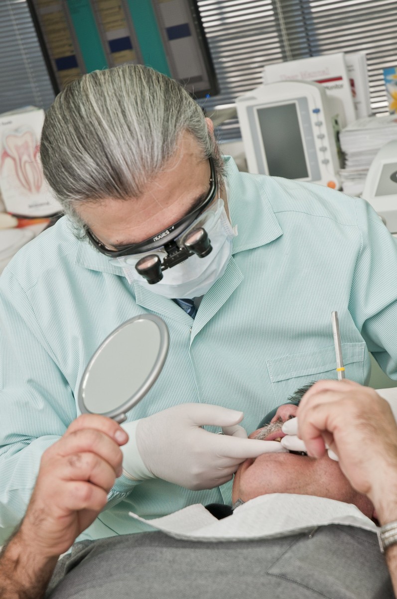 Person mit Mundschutz und Lupenbrille untersucht mit Spiegel und Instrument den Mund Person mit Mundschutz und Lupenbrille untersucht mit Spiegel und Instrument den Mund