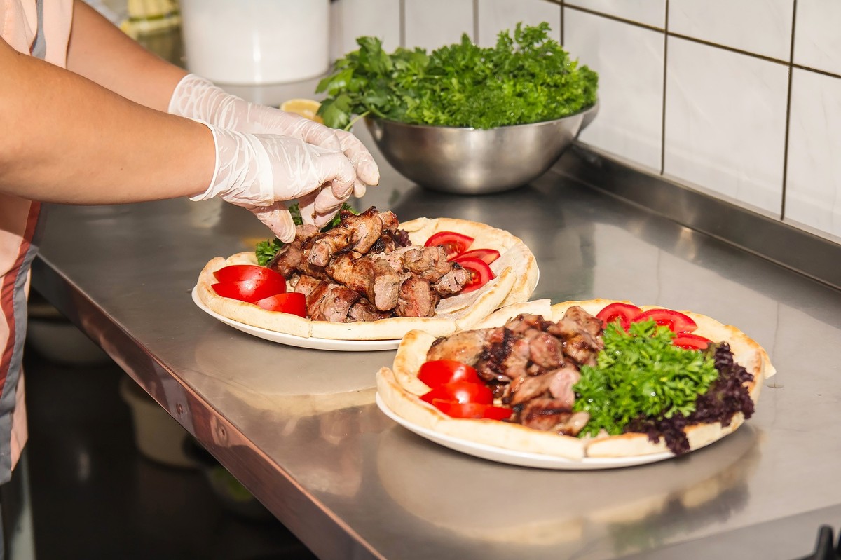 Hände in Handschuhen belegen Fladenbrote mit Fleisch, Tomaten und Salat Hände in Handschuhen belegen Fladenbrote mit Fleisch, Tomaten und Salat