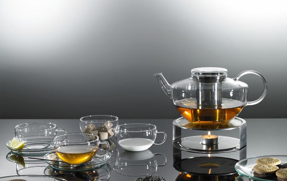 Teekanne mit Edelstahlfilter auf einem Stövchen, davor Teetassen, Milchkännchen, Zuckerschale und Gebäck