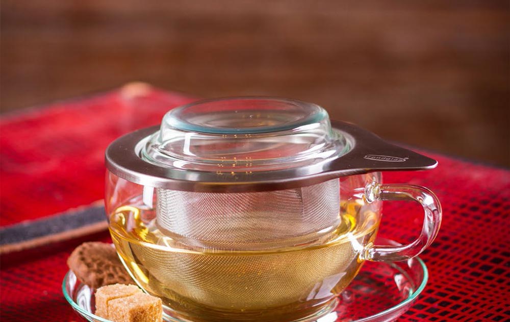 Teetasse mit Edelstahlsieb und Glasdeckel auf einer Untertasse mit Kandiszucker