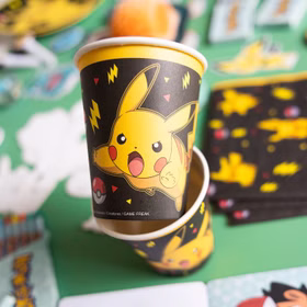 Pokémon Partybecher mit Pikachu-Motiv auf einem gedeckten Partytisch mit weiterer schwarz-gelber Pokémon Dekoration im Hintergrund.