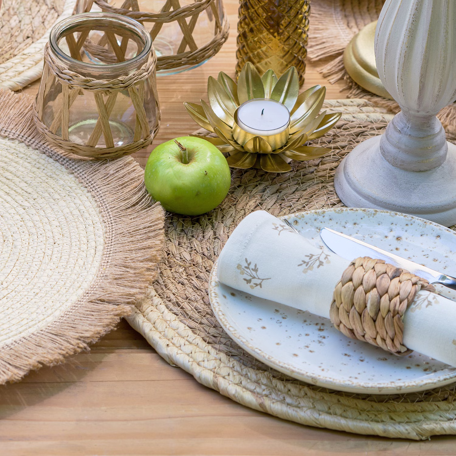 Boho-Tischdekoration mit rundem Stroh-Platzset, Kerzenhalter, Apfel, Geschirr und Naturaccessoires auf einem gedeckten Holztisch.