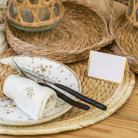 Naturfarbenes Platzset aus geflochtenem Stroh als Tischdeko mit Teller, Besteck, Serviette und Tischkärtchen im Boho-Stil.