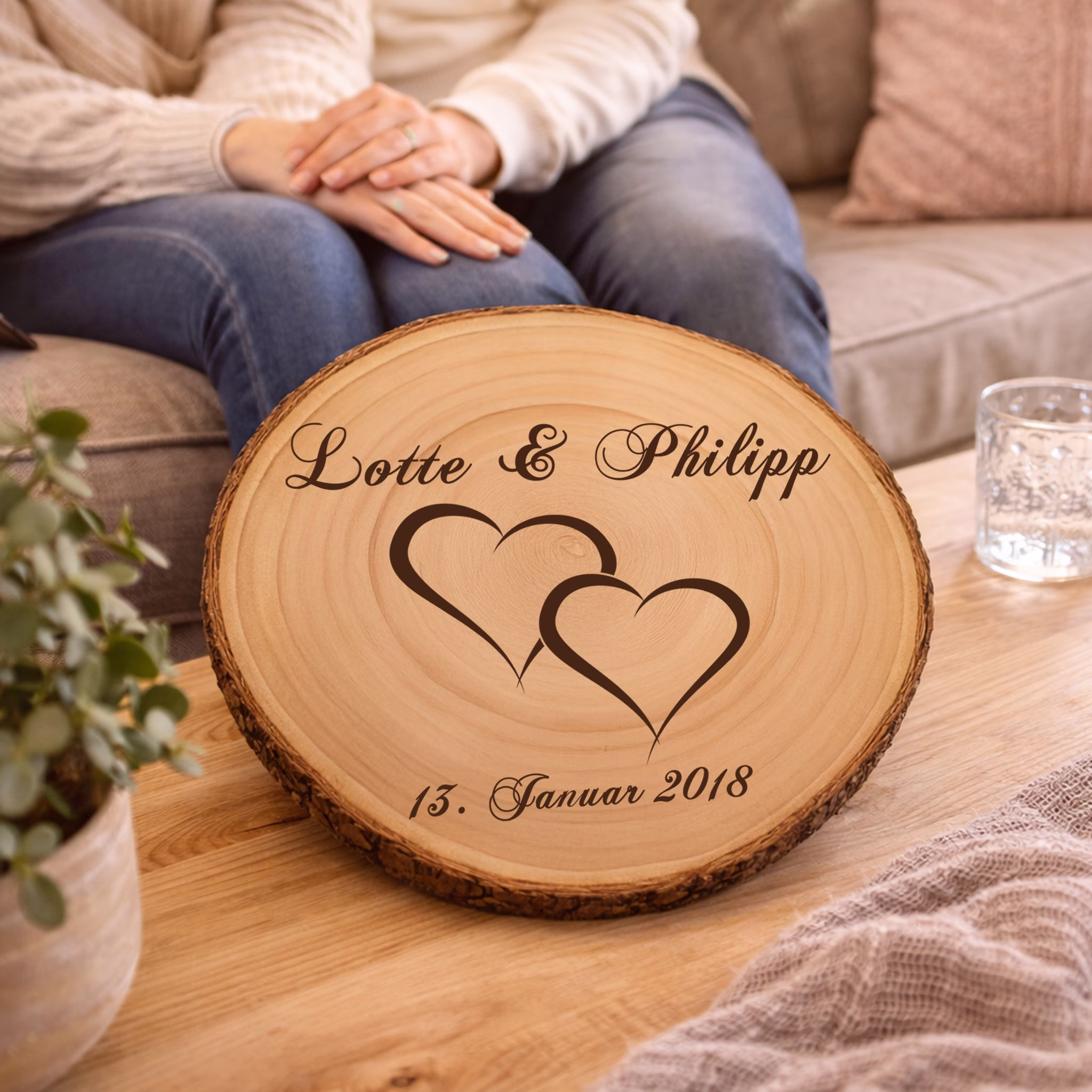 Baumscheibe mit Gravur Doppelherz Namen und Datum Hochzeit Jahrestag Holzscheibe Geschenkidee Brautpaar Hochzeitsgeschenk Personalisierte Baumscheibe mit Hochzeitsgravur auf einem Holztisch in einem gemütlichen Wohnzimmer, im Hintergrund sitzt ein Paar auf dem Sofa. Hinweis: KI generiertes Bild