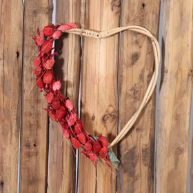 Großes Rattan-Herz hängt an einer Holzwand und ist dekorativ mit roten Trockenblumen geschmückt.
