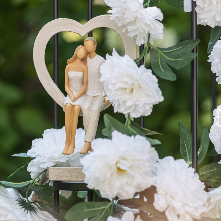 Brautpaar Figur Sitzend Liebe Weiß mit Herz Tischdeko Hochzeits-Deko Tisch-Deko Deko-Ideen Geschenkidee Braut & Bräutigam Brautpaar-Figur im Herzrahmen, dekoriert zwischen weißen Blumen im Garten.