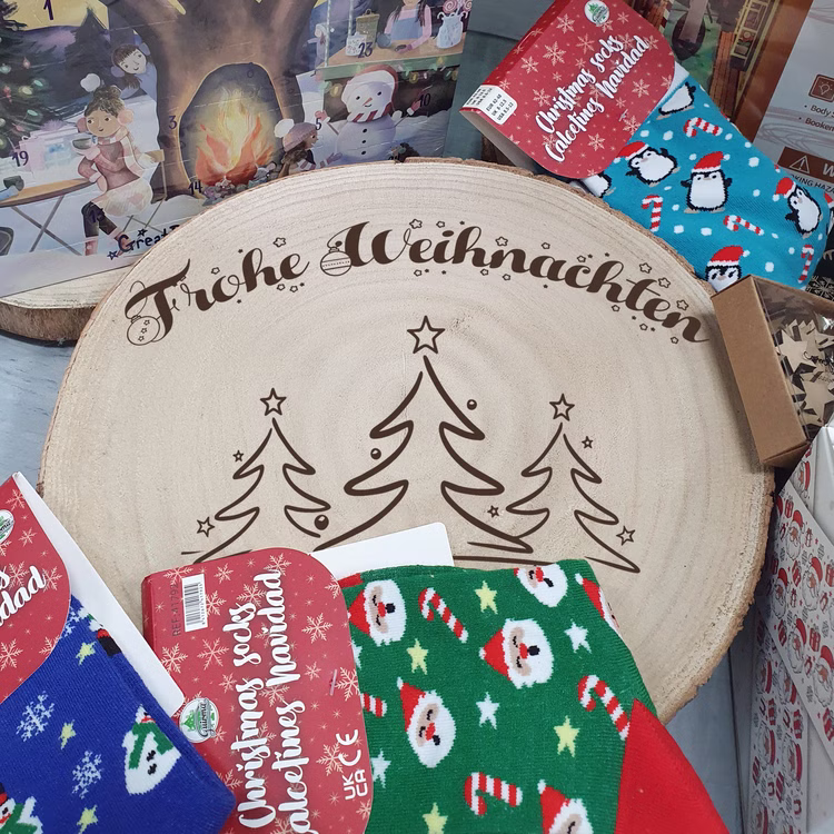 Baumscheibe 24 cm Frohe Weihnachten mit Tannenwald-Motiv Tisch-Deko Geschenk Dekoration Advent Weihnachtsstimmung festlich Adventszeit Basteln Geschenkidee Große gravierte Baumscheibe mit „Frohe Weihnachten“ und Tannenmotiv, umgeben von Weihnachtsartikeln wie Socken und Adventskalendern auf einem Tisch.