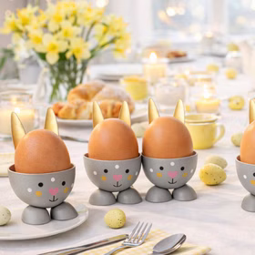 Mehrere graue Hasen-Eierbecher mit goldenen Ohren und braunen Hühnereiern auf einem hell gedeckten Oster-Frühstückstisch mit Blumen, Kerzen und Gebäck im Hintergrund.
