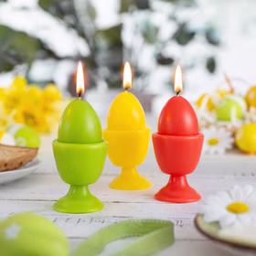 Drei brennende Eierkerzen in Rot, Grün und Gelb auf einem hell dekorierten Ostertisch mit Blumen im Hintergrund.