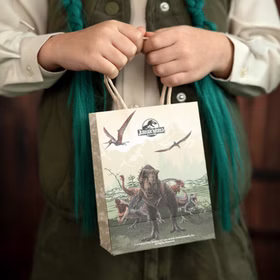 Jurassic World XXL Party-Set Dinosaurier Party-Deko 46-Teile T-Rex Geburtstag Saurier Lizenz-Party-Zubehör gedeckter Tisch Tisch-Deko Mottoparty Kindergeburtstag Geburtstag Kind hält eine Jurassic World Geschenktüte mit Dinosaurier-Motiv vor dem Körper.