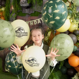 Jurassic World XXL Party-Set Dinosaurier Party-Deko 46-Teile T-Rex Geburtstag Saurier Lizenz-Party-Zubehör gedeckter Tisch Tisch-Deko Mottoparty Kindergeburtstag Geburtstag Kind spielt mit mehreren Jurassic World Ballons vor einer Dino-Geburtstagskulisse in Grün.