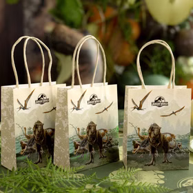 Jurassic World XXL Party-Set Dinosaurier Party-Deko 46-Teile T-Rex Geburtstag Saurier Lizenz-Party-Zubehör gedeckter Tisch Tisch-Deko Mottoparty Kindergeburtstag Geburtstag Drei Jurassic World Geschenktüten auf dekoriertem Partytisch mit grüner Dino-Deko im Hintergrund.