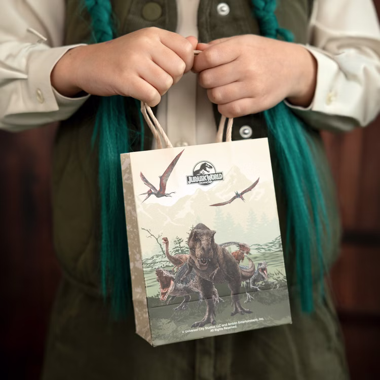Jurassic World XXL Party-Set Dinosaurier Party-Deko 46-Teile T-Rex Geburtstag Saurier Lizenz-Party-Zubehör gedeckter Tisch Tisch-Deko Mottoparty Kindergeburtstag Geburtstag Kind hält eine Jurassic World Geschenktüte mit Dinosaurier-Motiv vor dem Körper.