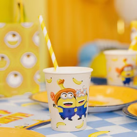 Minions Partybecher Tischdekoration Minions Partybecher mit gelb-weißem Trinkhalm auf einer blau karierten Geburtstagstafel mit Tellern und gelber Dekoration.