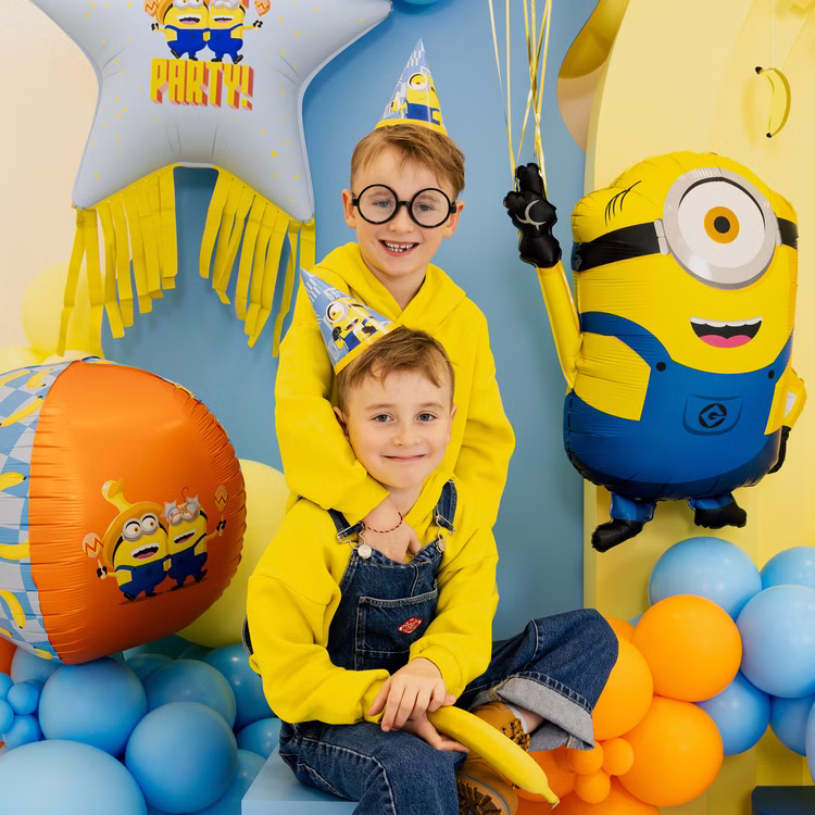 Minions Geburtstag Kinder Partydekoration Zwei Jungen mit Minions Partyhütchen zwischen gelben, blauen und orangefarbenen Ballons und großen Minions Dekoelementen.
