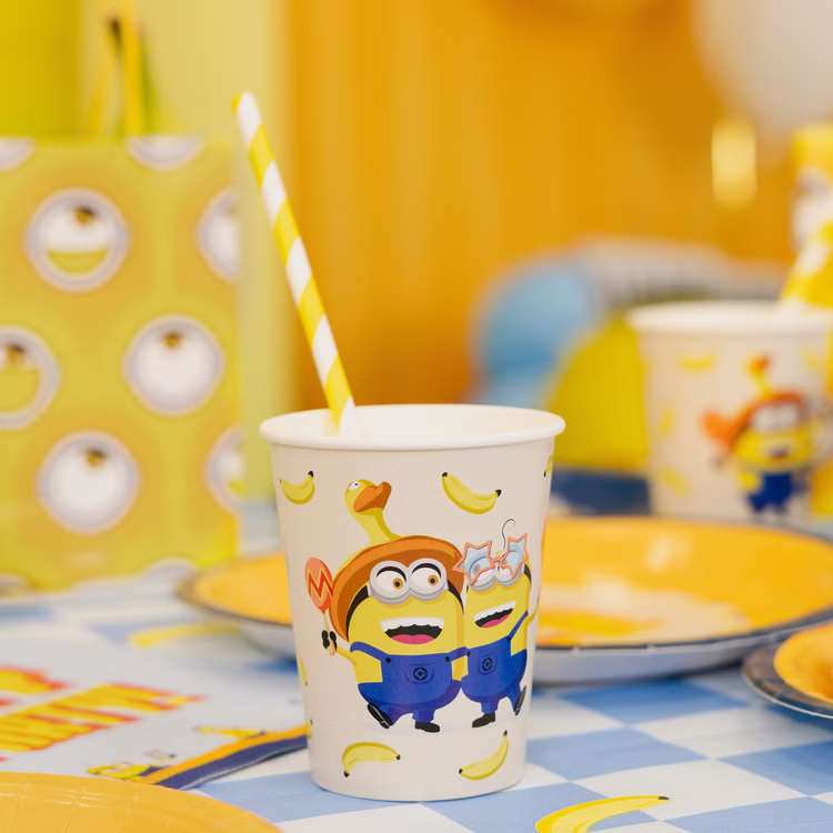 Minions Partybecher Tischdekoration Minions Partybecher mit gelb-weißem Trinkhalm auf einer blau karierten Geburtstagstafel mit Tellern und gelber Dekoration.