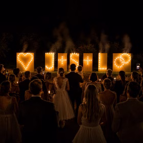 Lichterbilder Lichterbild Brennende Buchstaben Feuerwerk Geschenkidee Geburtstag Goldene Hochzeit Jubiläum Brautpaar mit Kind betrachtet gemeinsam ein brennendes Hochzeits-Lichterbild vor einer festlichen Hochzeitsgesellschaft bei Nacht.