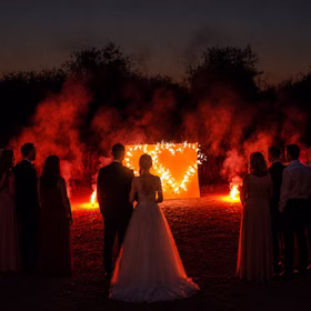 Hochzeitsgesellschaft mit Braut und Bräutigam betrachten das brennende Lichterbild Motiv Doppelherz mit Bengalfeuer bei Abenddämmerung.