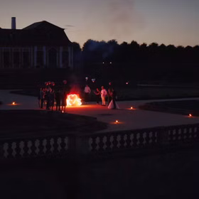 Hochzeitsgesellschaft mit Ballons und Fackeln um ein brennendes Herz vor einem Schloss bei Dämmerung.