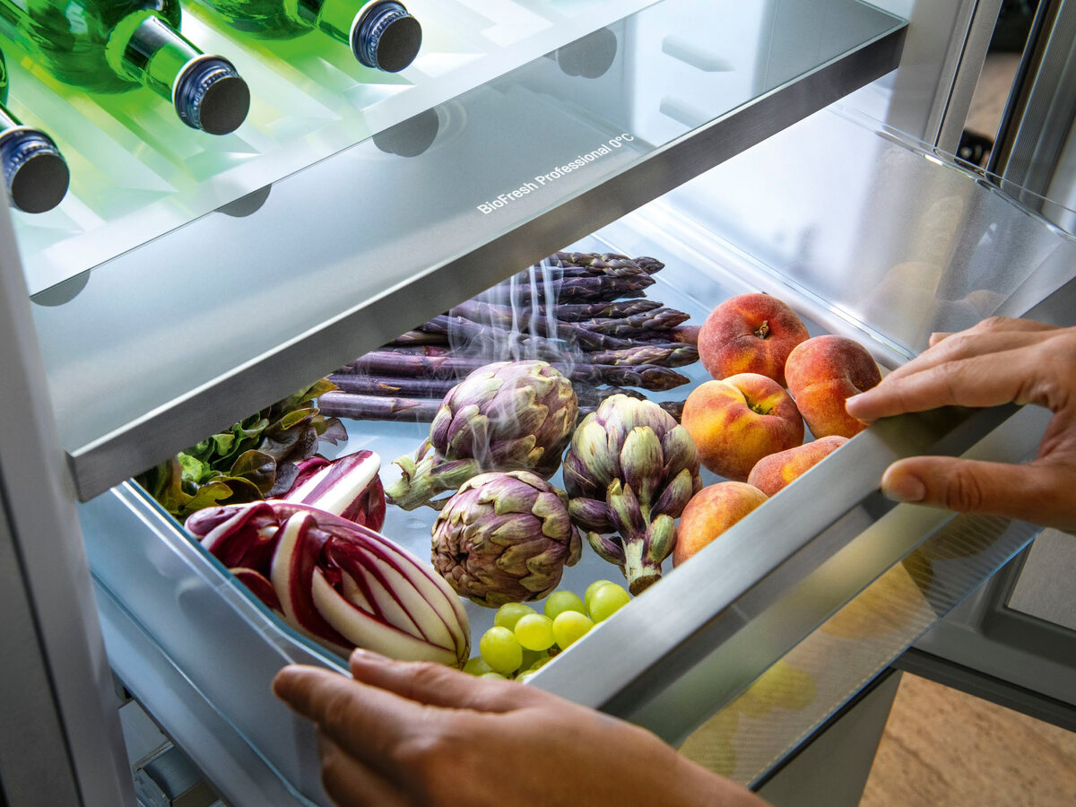 Geöffnete BioFresh-Schublade mit Artischocken, Pfirsichen, Spargel und Radicchio, darüber feiner Nebel; zwei Hände greifen in die Lade.
