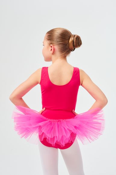 Ein junges Mädchen in einem rosa Ballettbody und Tutu steht mit den Händen in den Hüften und blickt von der Kamera weg.