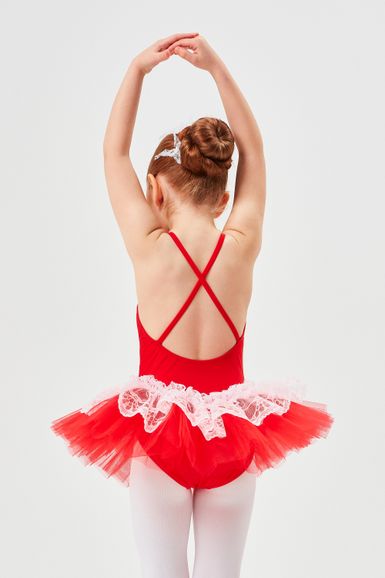 Ein junges Mädchen in einem roten Tutu und weißen Strumpfhosen steht mit erhobenen Armen und blickt von der Kamera weg.