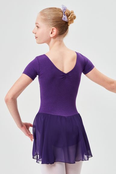 Ein junges Mädchen in einem lila Ballettbody und Rock posiert in einer Balletthaltung und blickt von der Kamera weg.