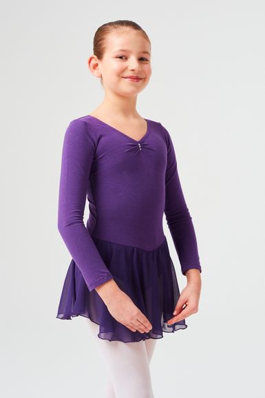 Ein junges Mädchen in einem lila Langarm-Trikot und Rock steht lächelnd.