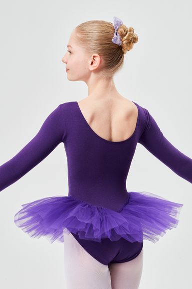Mädchen in einem lila Ballettbody und Tutu steht mit leicht ausgestreckten Armen da und blickt von der Kamera weg.