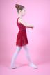 Junges Mädchen in einem roten Ballettbody und Rock posiert in Ballettposition vor einem schlichten rosa Hintergrund. 1