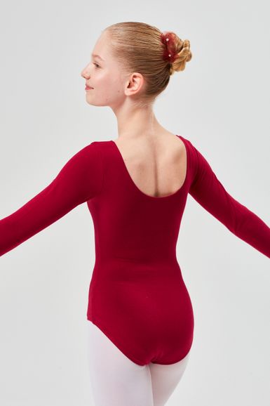Eine junge Balletttänzerin in einem roten Ballettbody und weißen Strumpfhosen steht mit ausgestreckten Armen da und blickt leicht weg.