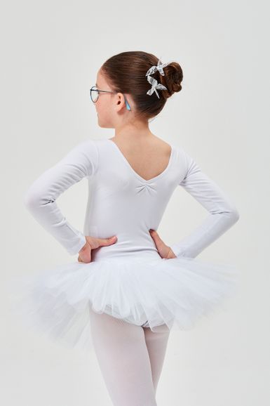 Ein junges Mädchen in einem weißen Ballett-Outfit  steht mit den Händen in den Hüften und blickt von der Kamera weg.