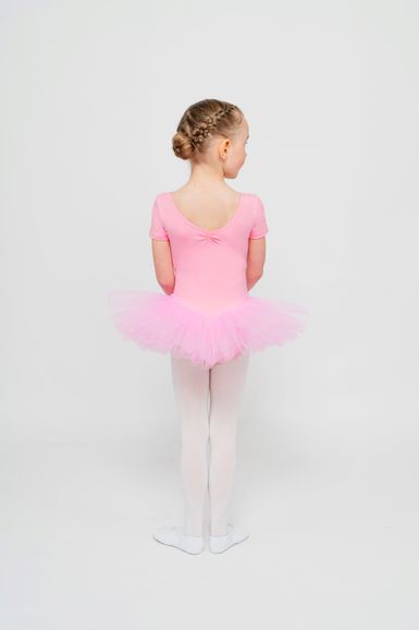 Ein kleines Kind in einem rosa Ballett Trikot und Tutu steht mit dem Rücken zu einem schlichten weißen Hintergrund.