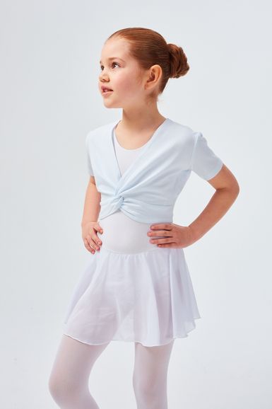 Ein junges Mädchen in einem weißen Ballett-Outfit und Strumpfhosen steht mit den Händen in den Hüften und blickt zur Seite.