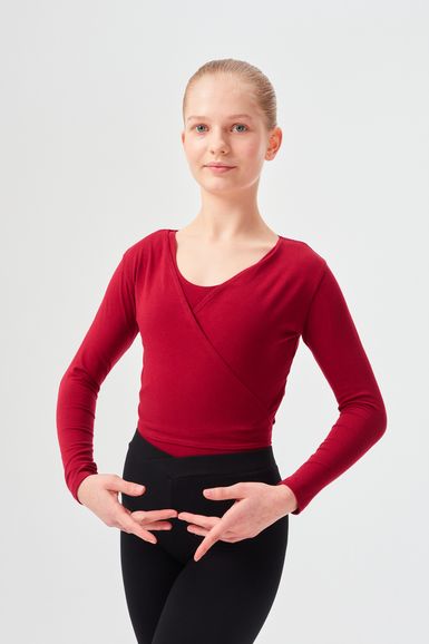Junge Person in rotem Langarmshirt und schwarzer Hose posiert mit den Händen in Ballettposition vor schlichtem Hintergrund.