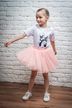Ein junges Mädchen in einem weißen T-Shirt und einem rosa Tutu steht auf einem Holzboden vor einer weißen Ziegelwand und posiert mit ausgestreckten Armen. 4