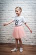 Ein junges Mädchen in einem weißen Ballett-T-Shirt und einem rosa Tutu steht mit ausgestreckten Armen vor einer weißen Backsteinwand. 3