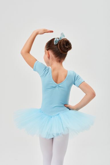 Ein junges Mädchen in einem blauen Ballett-Outfit steht in einer Tanzpose mit erhobenem Arm und dem Rücken zur Kamera.