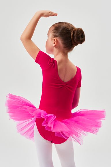 Ein junges Mädchen in einem rosa Ballett Trikot und Tutu steht mit erhobenem Arm da und blickt von der Kamera weg.