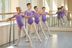 Vier junge Mädchen in lila Ballettbodys üben Ballett an einer Stange in einem Tanzstudio mit großen Fenstern. 3