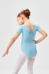 Ein junges Mädchen in einem hellblauen Ballett Trikot und weißen Strumpfhosen steht mit einer Hand auf der Hüfte und blickt von der Kamera weg. 2
