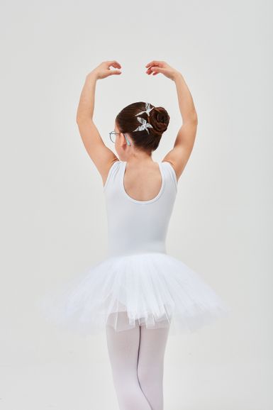 Ein junges Mädchen in einem weißen Ballett-Outfit steht mit über dem Kopf erhobenen Armen da und blickt von der Kamera weg.