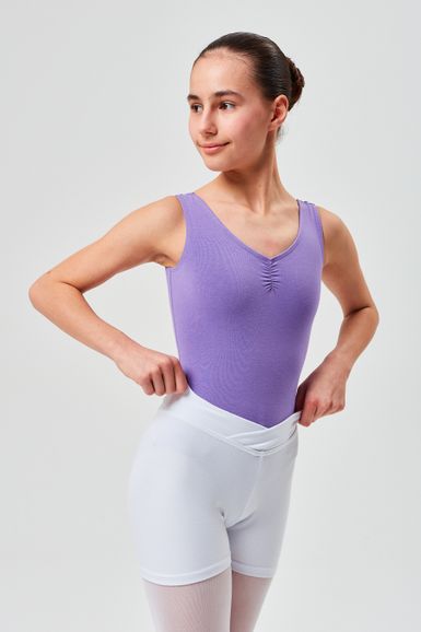 Eine junge Balletttänzerin in einem lila Ballett Trikot und weißen Shorts steht mit den Händen in den Hüften da und schaut zur Seite.