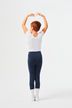 Ein Kind in einem weißen Oberteil und marineblauen Leggings steht in einer Ballettpose mit erhobenen Armen und blickt von der Kamera weg.