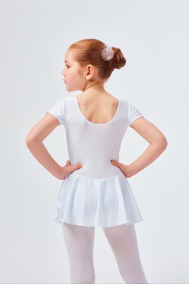 Ein junges Mädchen in einem weißen Ballett-Outfit steht mit den Händen in den Hüften und blickt von der Kamera weg.