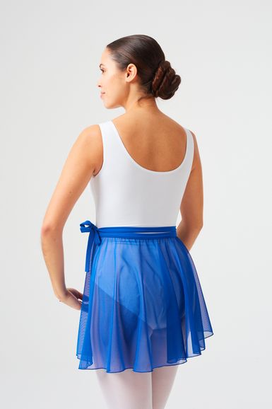 Eine Frau in einem weißen Trikot und einem blauen Ballettrock steht mit dem Rücken zur Wand in einem Studio mit schlichtem Hintergrund.