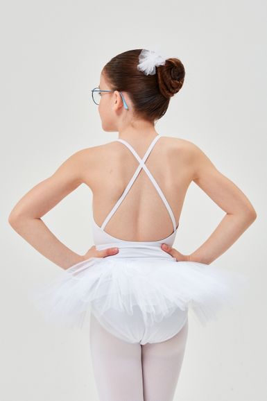 Ein junges Mädchen in einem weißen Ballett-Outfit  steht mit den Händen in den Hüften und dem Rücken zu ihr.