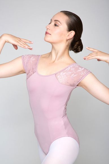 Eine Balletttänzerin in einem hellrosa Trikot und weißen Strumpfhosen posiert mit erhobenen Armen und blickt zur Seite.