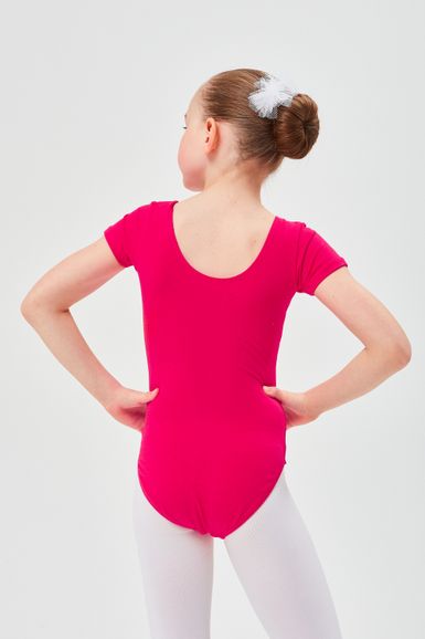 Ein junges Mädchen in einem rosa Ballettbody und weißen Strumpfhosen steht mit den Händen in den Hüften und dem Rücken zu.