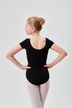 Eine junge Balletttänzerin in einem schwarzen Ballettbody und rosa Strumpfhosen steht mit den Händen in den Hüften und dem Rücken zu. 2